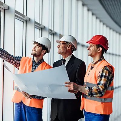 Immagine copertina corso online. Due lavoratori vestiti da tecnici con gilet e caschi di sicurezza e un uomo distinto con caso sicurezza controllano la planimetria dell'edificio